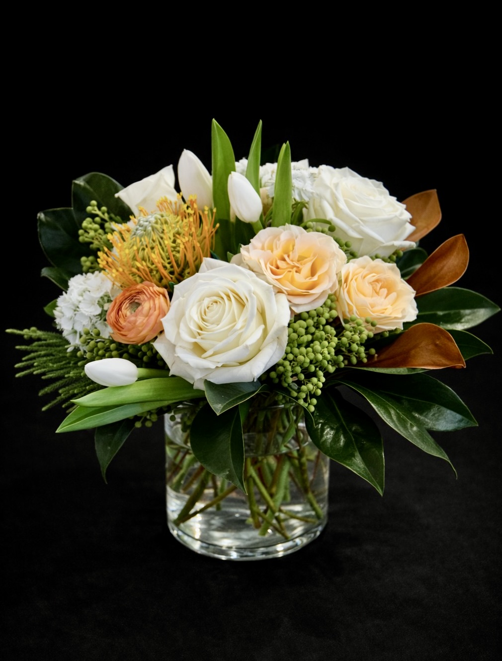 Luxury seasonal floral arrangement with white roses, peach garden roses, tulips, ranunculus, and pincushion protea in a glass vase on a black background by Vail Colorado florist A Secret Garden